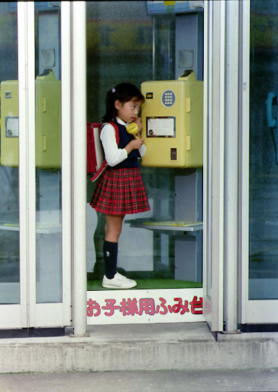 電話ボックスのプッシュ式公衆電話で電話をする小学生の女の子の写真
